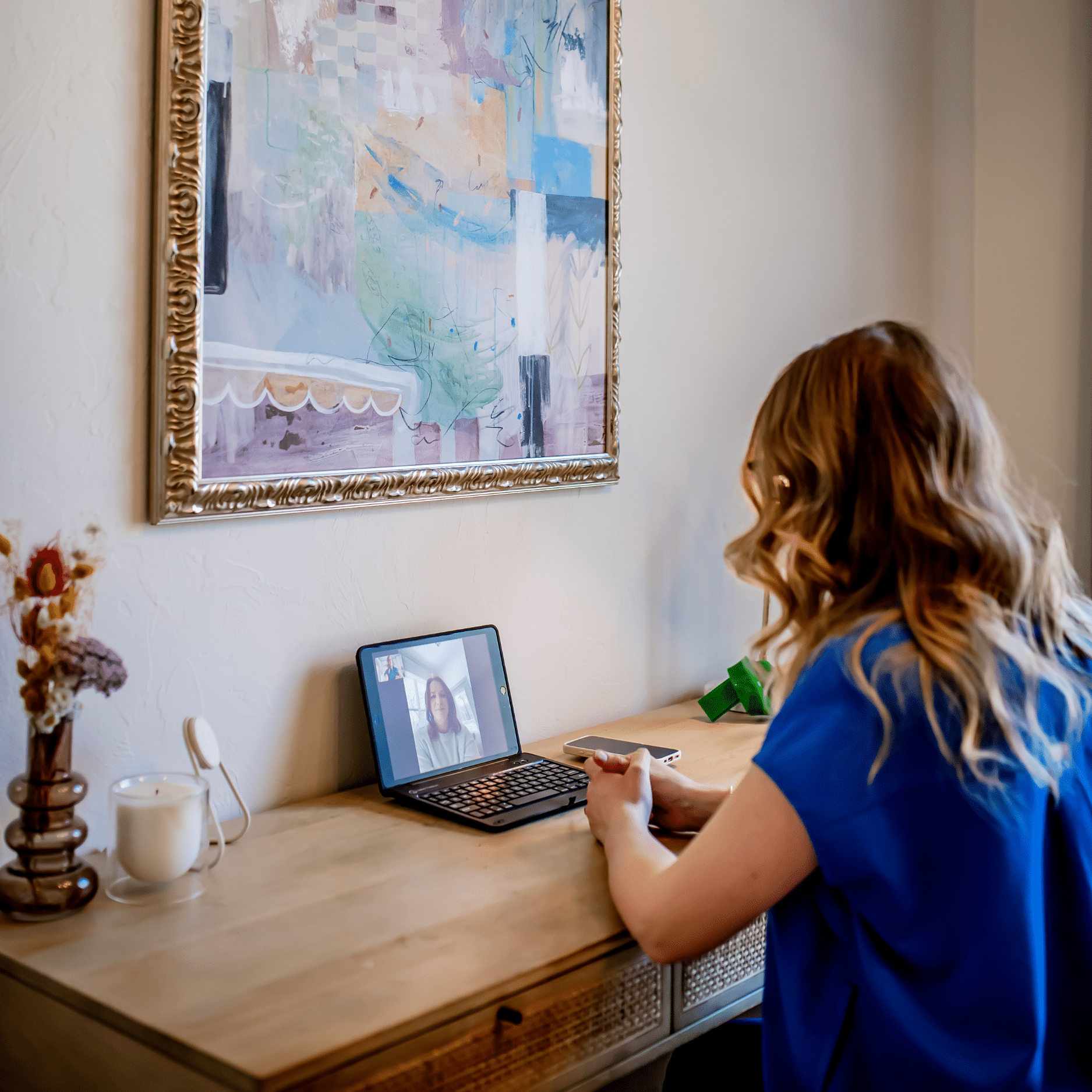 Professional woman with reddish-brown hair wearing a blue top sitting at a desk during a virtual meeting on a laptop, with an abstract framed artwork hanging on the wall behind her